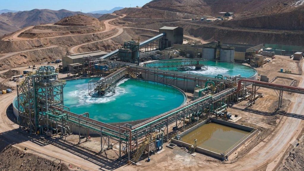 Vista aérea panorámica de una planta de procesamiento minero en el norte de Chile, mostrando grandes piscinas circulares de espesamiento con agua de procesos de color turquesa, rodeadas de una compleja red de tuberías, estructuras metálicas industriales y caminos de tierra en un entorno cordillerano árido.